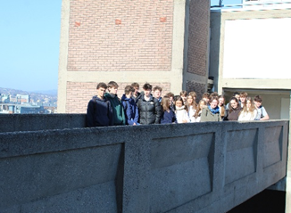 Photograph of a group of young people standing closely together on a concrete balcony or bridge with a brick building and cityscape in the background.