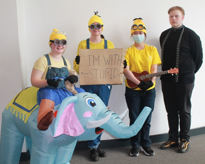 Four people dressed in costumes stand against a plain wall, with three wearing yellow Minion outfits and one in black clothing. One person holds a cardboard sign reading "I'M WITH STUPID," while another wears an inflatable elephant costume.