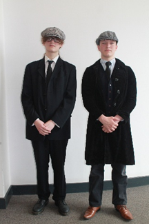 Photograph of two people standing side by side against a plain wall, both dressed in formal black suits, white shirts, black ties, and patterned flat caps. One wears brown shoes and a velvet jacket, while the other wears black shoes and a standard suit jacket, both with hands clasped in front.