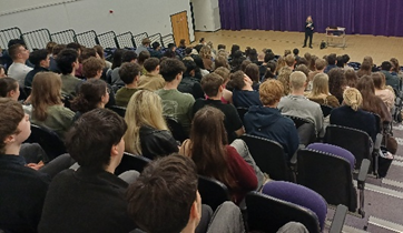 Photograph of a large group of students seated in an auditorium facing a speaker on stage. The setting features tiered seating with black and purple chairs, a wooden floor, and a purple curtain backdrop,