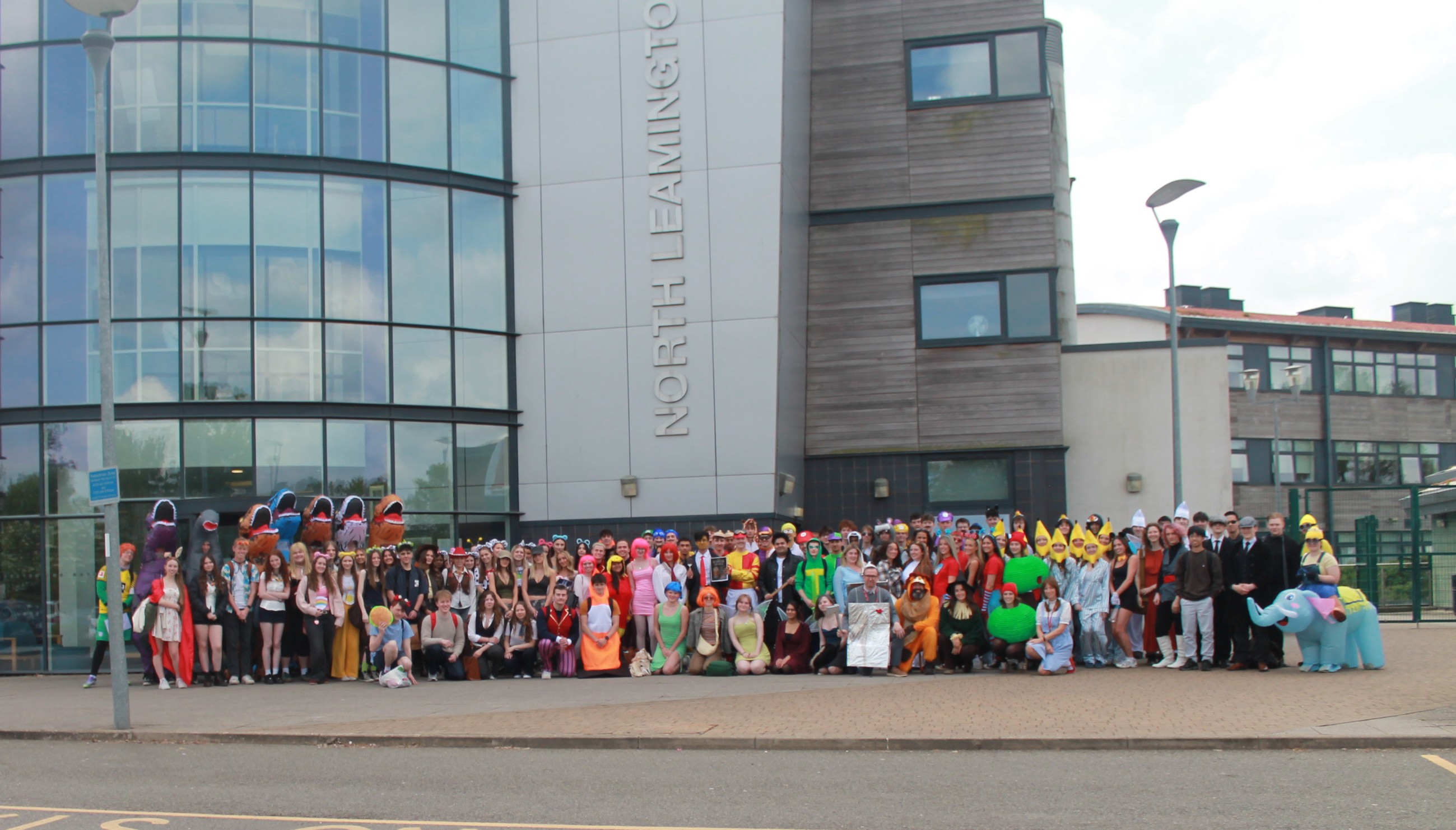 Photograph showing a large group of people dressed in various colourful costumes standing outside a modern building labelled "North Leamington." The scene captures a festive or themed event with diverse outfits, including characters and animals,