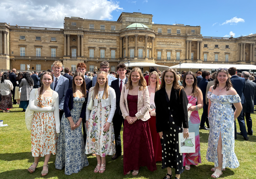 Photograph showing a group of young people dressed formally standing on grass in front of a large historic building under a partly cloudy sky at Buckingham Palace in a formal event or gathering, with individuals wearing suits and dresses in various colours and patterns, and a crowd visible in the background.
