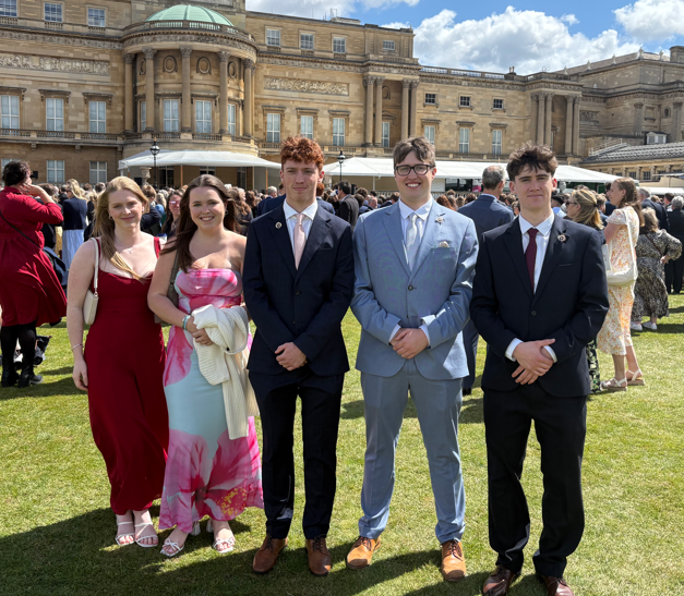 Photograph of a formal outdoor gathering on a lawn in front of a large historic building under a partly cloudy sky. Five young adults dressed in formal attire stand in the foreground, with a crowd of similarly dressed people mingling in the background.