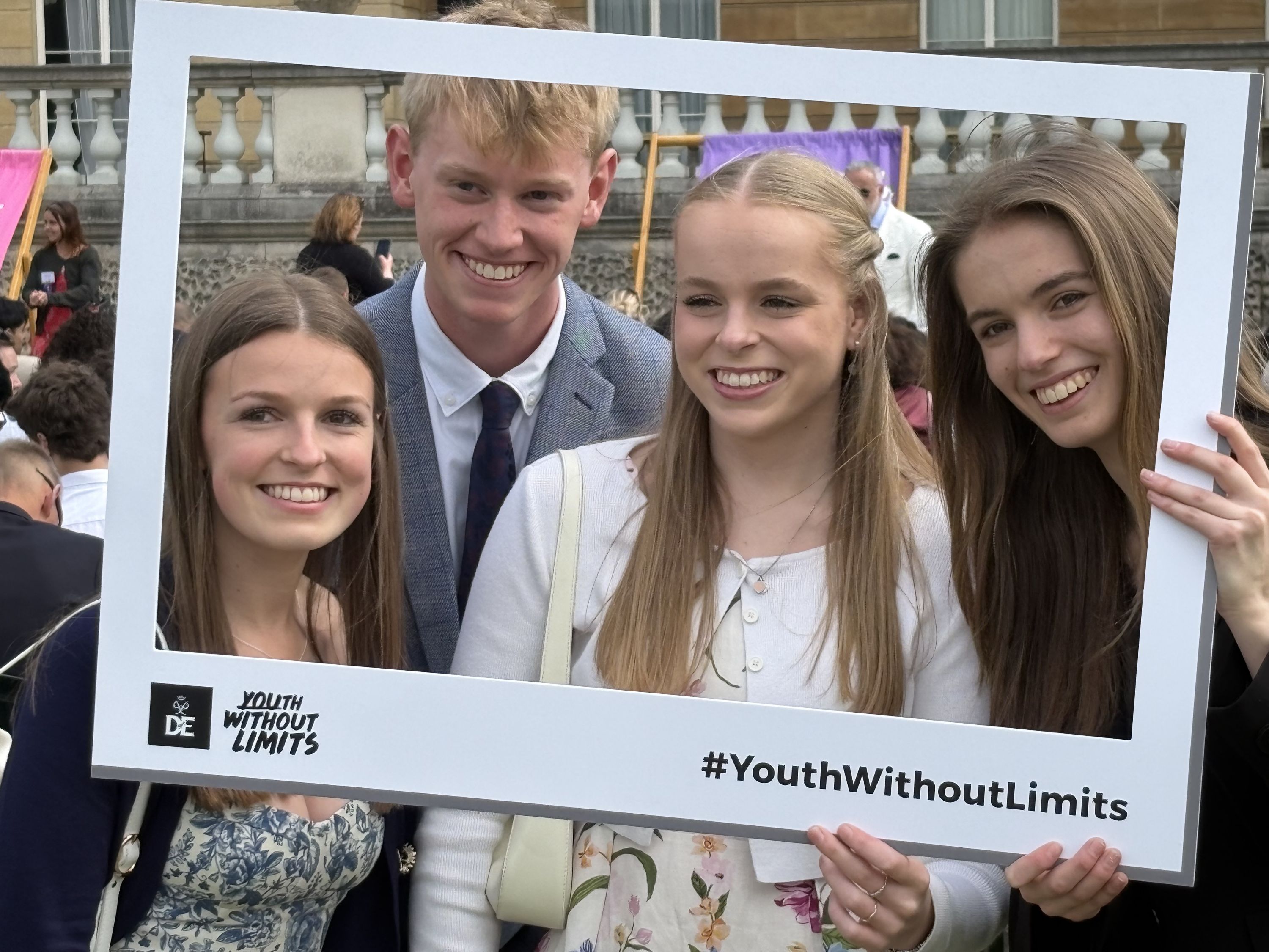 Photograph of four young people posing outdoors while holding a large white rectangular frame with "Youth Without Limits" logo and hashtag #YouthWithoutLimits at Buckingham Palace