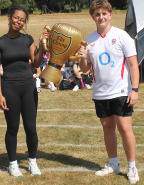 Photograph of two young people standing on a grassy field holding a large inflatable gold trophy labelled "The Champ." The person on the left wears black athletic clothing and glasses, while the person on the right wears a white sports jersey with "O2" and black shorts, with other people and trees visible in the background.