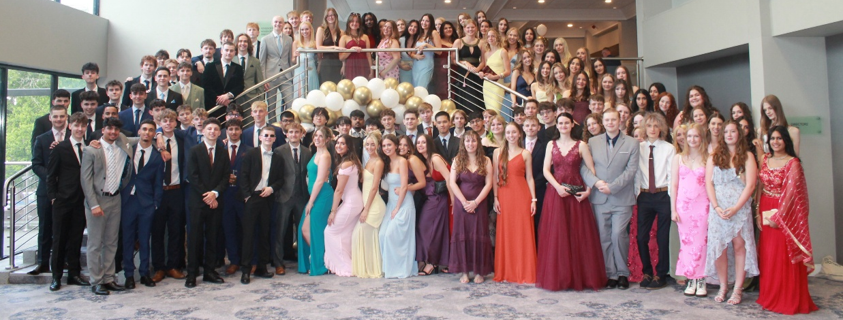 Photograph of a large group of young people dressed formally, posing indoors on and around a staircase decorated with gold and white balloons. The setting suggests a formal event such as a prom or graduation, with individuals wearing suits and a variety of colourful evening dresses.