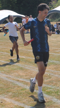 Photograph of a relay race on a grass track with runners passing a red baton. Foreground runner wears a dark blue sports kit, while background runners and spectators are visible under white tents.