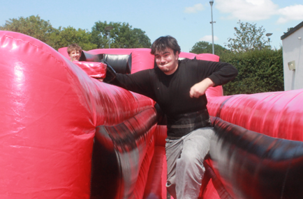 Photograph showing a young man running through a red and black inflatable obstacle course outdoors on a sunny day. Another person is visible in the background, partially obscured by the inflatable structure and greenery