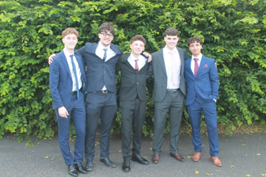 Photograph of five young men dressed in formal suits standing side by side outdoors with arms around each other, posing in front of dense green foliage. Each person wears a different coloured suit and tie, suggesting a celebratory or special occasion such as prom or a formal event.