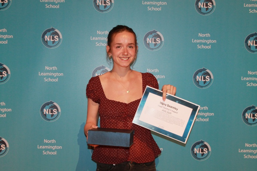 a young woman standing against a North Leamington School branded backdrop, smiling while holding a certificate and a blue box. The setting suggests an award or recognition event, with the certificate prominently displayed and the backdrop featuring repeated school logos and name in white on a teal background.