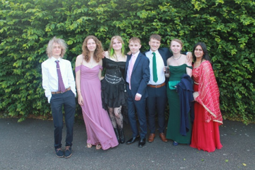 Photograph of seven young adults dressed formally, standing side by side outdoors against dense green foliage. Attire includes suits and elegant dresses in various colours, with one person wearing a traditional red and gold outfit, suggesting a celebratory event like prom or a formal gathering.