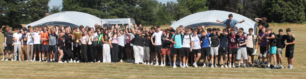 Photograph showing a large group of people standing on grass in front of two white tents, with many raising their hands and smiling. The scene suggests a social or community event held outdoors on a sunny day.