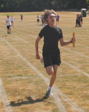 Photograph of a relay race on a grass track with multiple runners competing. Foreground features a young male athlete in black sportswear holding a yellow baton, running towards the camera with other participants and spectators visible in the background
