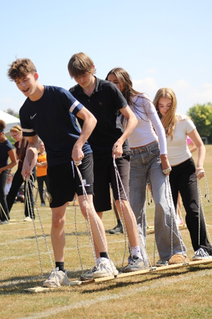 Photograph showing a group of young people participating in an outdoor team-building activity involving ropes. Participants stand closely on grass, focused on coordinating movements, with clear blue sky and trees in background.