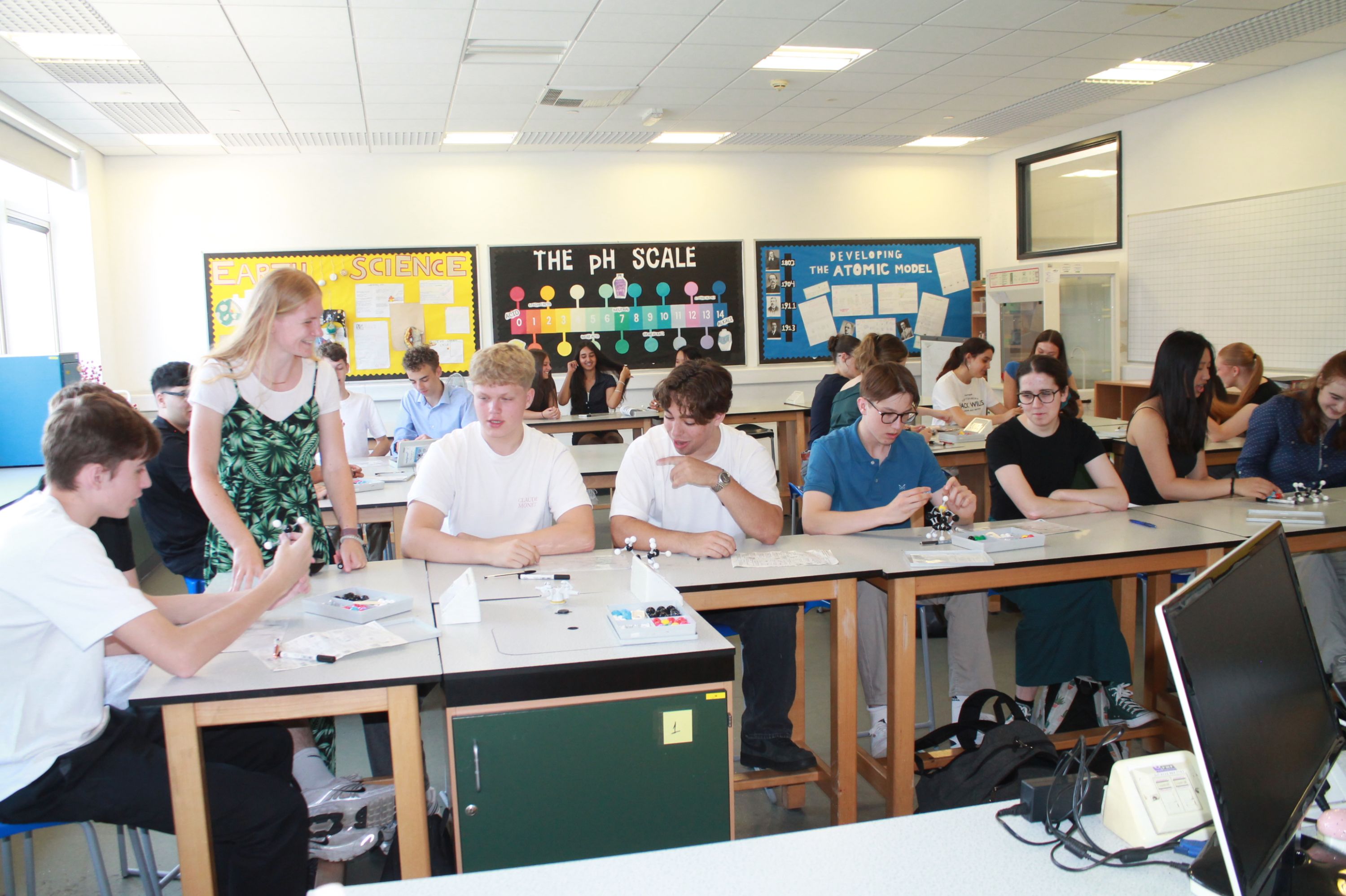Photograph of a classroom with students seated at lab tables engaged in a science activity, with a teacher assisting one student. Colourful educational posters on pH scale and atomic model are visible on walls, indicating a chemistry lesson setting.