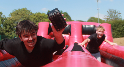 Photograph of two young men participating in an outdoor inflatable obstacle course, with one man reaching forward holding a black object. Bright red and black inflatable structure contrasts with green trees and clear blue sky in background, capturing a moment of active competition and enjoyment.