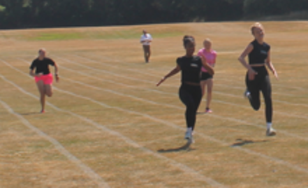 Photograph of four people sprinting on a grass running track marked with white lanes during daytime. Two runners in black are leading, with one in pink following behind, set against a background of trees and open field.