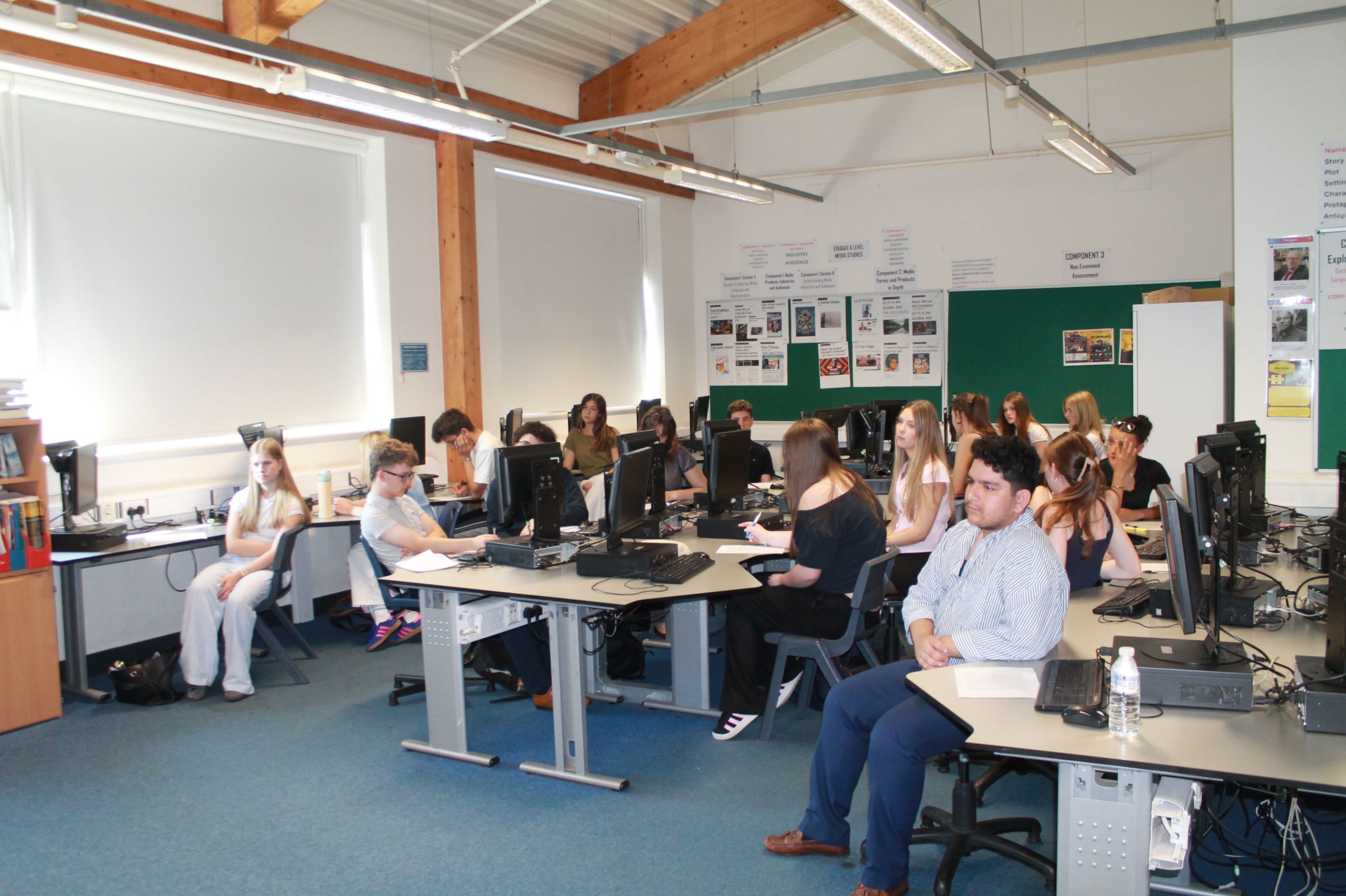 Photograph of a classroom with multiple students seated at desks equipped with desktop computers, engaged in various activities. The room features large windows with blinds, exposed wooden beams, and walls decorated with posters and notices, indicating an educational  environment