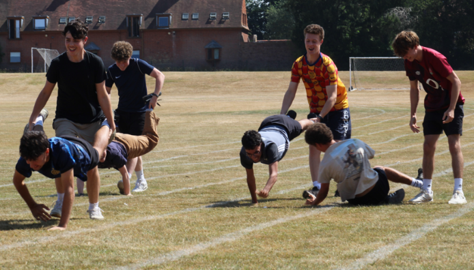Photograph of a group of young people participating in a wheelbarrow race on a grass field marked with white lines. Participants are paired, with one person holding the legs of another who is walking on their hands, showing active engagement and teamwork in an outdoor sports or recreational event.
