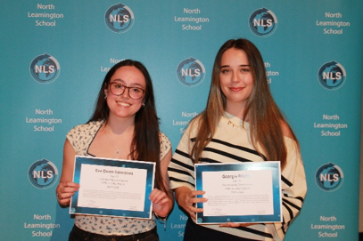wo young women standing in front of a blue backdrop with repeated North Leamington School logos, each holding a certificate. Both are smiling, with one wearing glasses and a patterned top, and the other in a striped sweater, indicating an award or achievement recognition event.