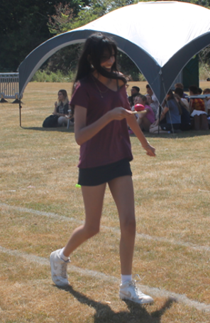 Photograph of a young woman walking on a grassy field during an outdoor event, wearing a maroon t-shirt, black shorts, white sneakers, and a face mask. A white and blue canopy with seated people is visible in the background