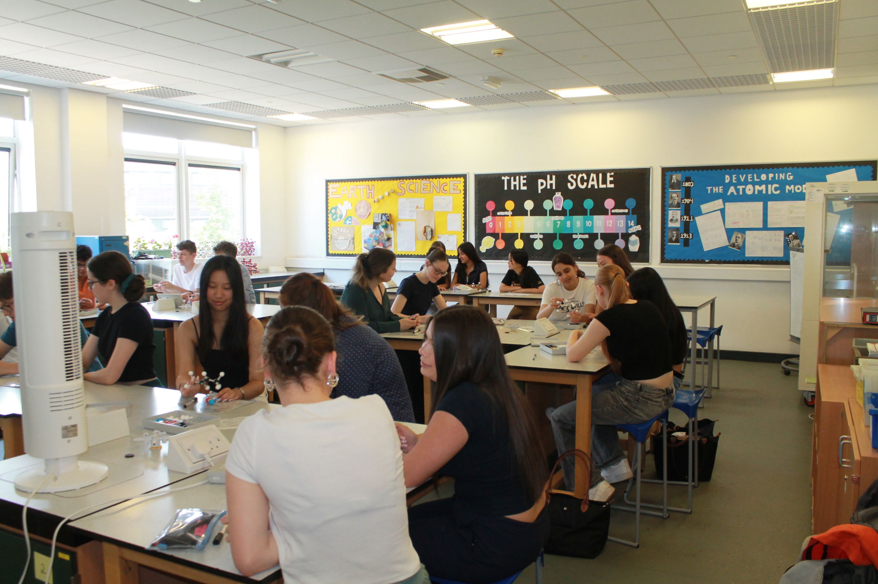 Photograph of a science classroom with students seated at lab tables engaged in experiments or discussions. Colourful educational posters on pH scale and atomic model displayed on walls, with natural light from large windows illuminating the room.