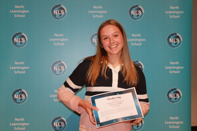a smiling young girl holding a certificate in front of a blue backdrop featuring repeated North Leamington School logos and text. The setting suggests a school award or recognition event celebrating student achievement.