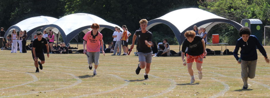 Photograph of five young people sprinting on marked grass lanes during an outdoor race event. White canopy tents and spectators are visible in background, indicating a casual sports day or community gathering.