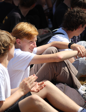 Photograph of a group of young people sitting outdoors, engaging in conversation and clapping. The scene highlights casual clothing, natural light, and a relaxed social atmosphere.