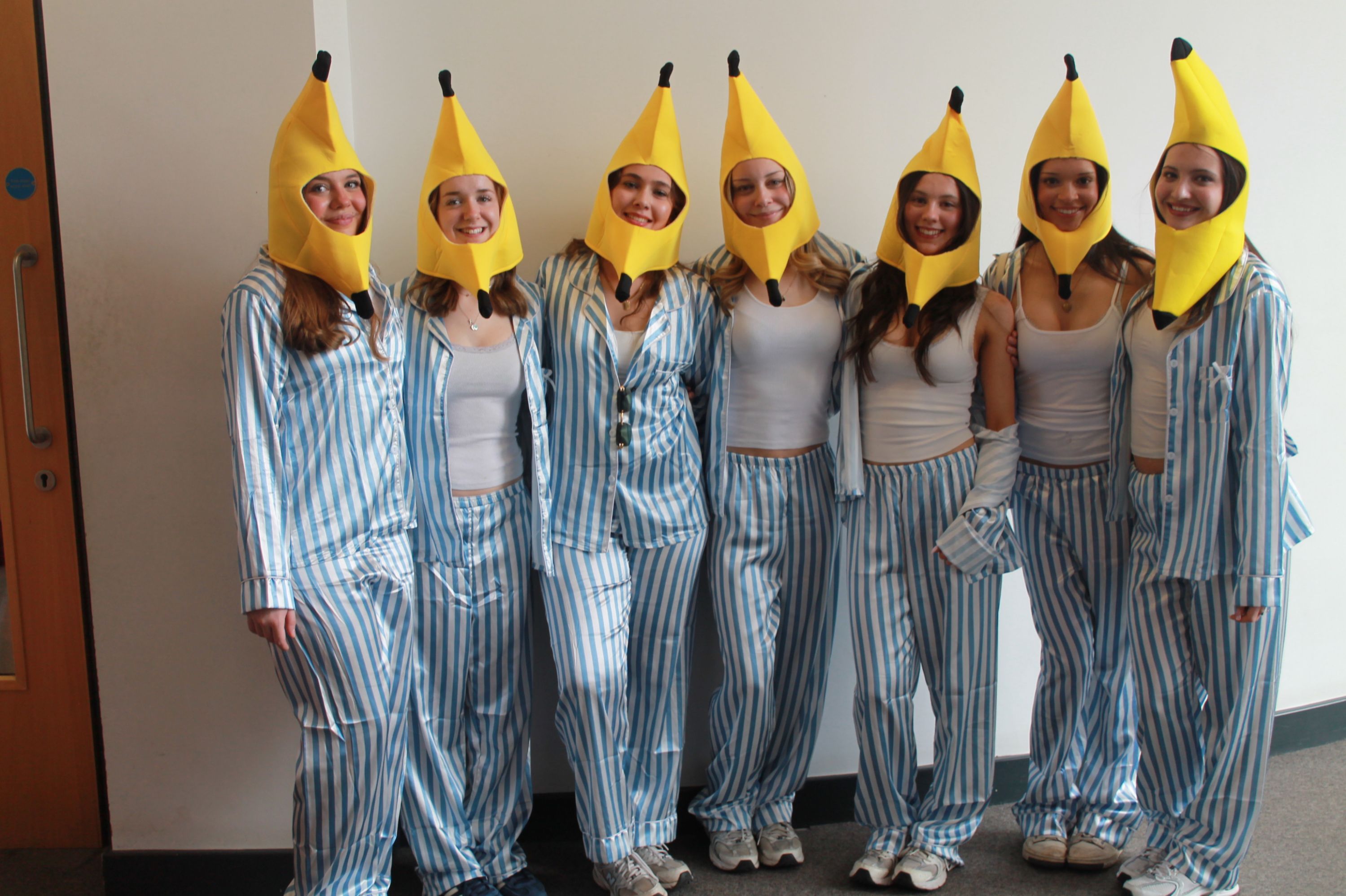 Photograph of seven people standing side by side wearing matching blue and white striped pyjamas and large yellow banana hat