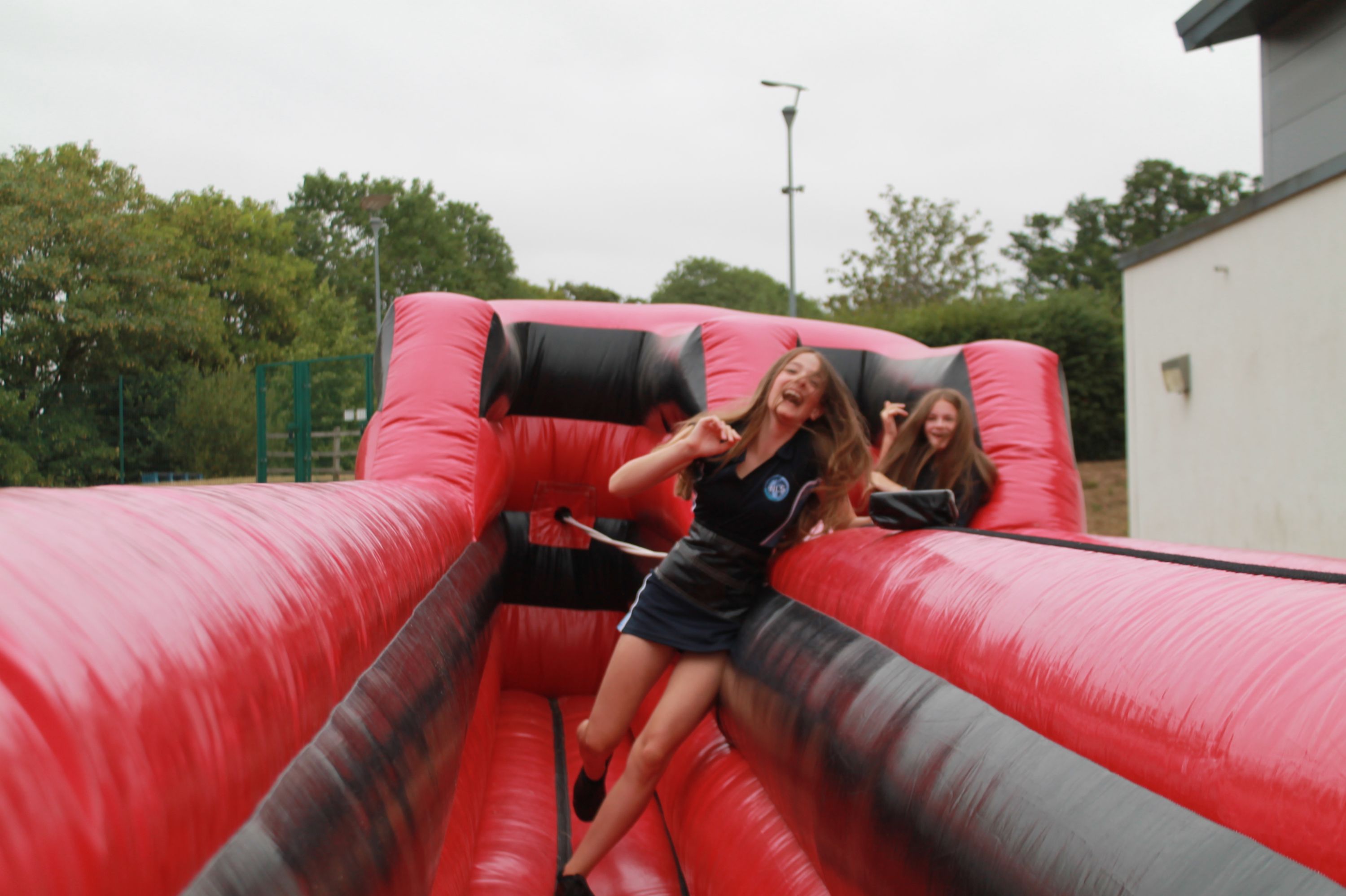 Photograph showing two young women playing on a large red and black inflatable game outdoors near a building and trees. The women appear joyful and engaged, with one sliding down the inflatable while the other watches from behind.