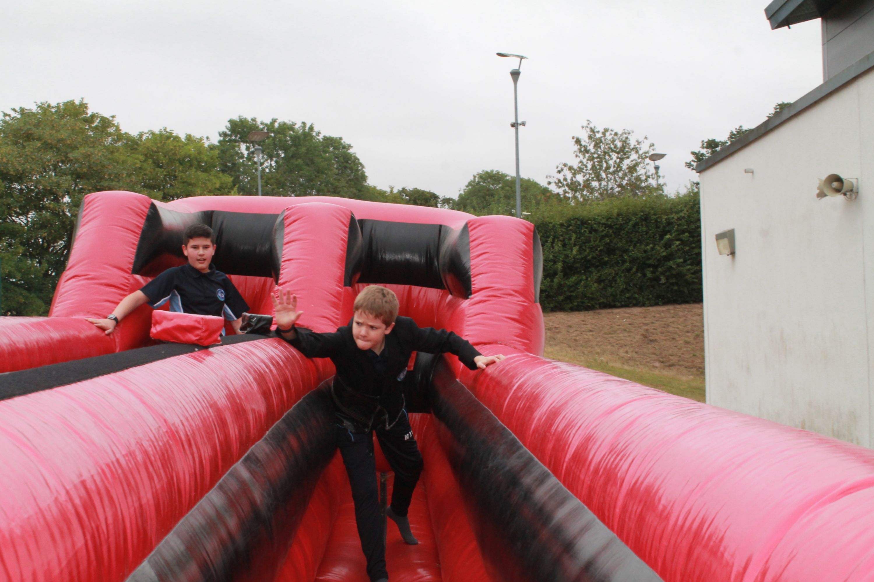 Photograph showing two boys playing on a large red and black inflatable obstacle course outdoors. One boy is climbing up while the other is seated at the top, with trees and a building visible in the background under an overcast sky.
