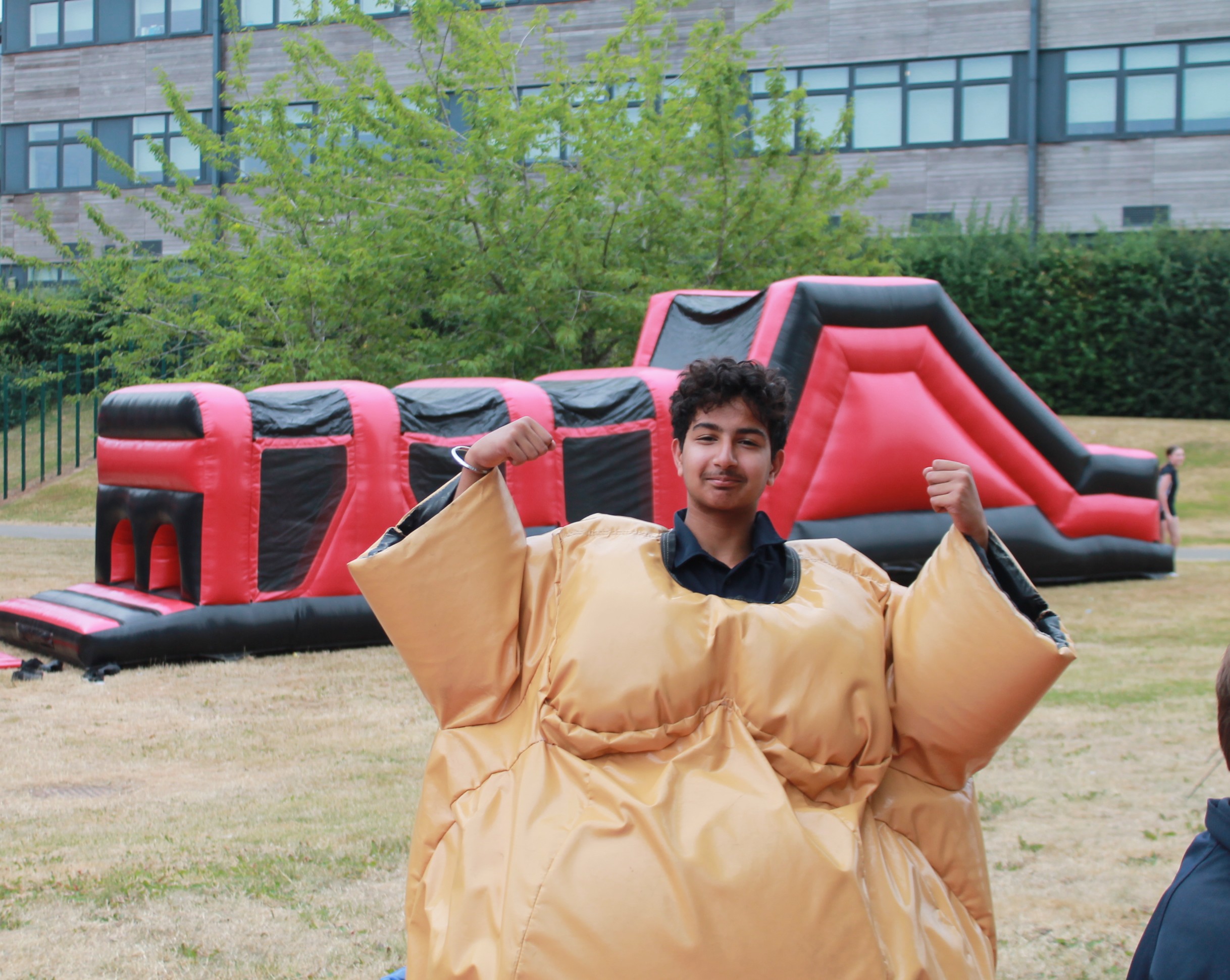 Photograph of a person wearing an oversized sumo wrestling suit outdoors on grass with a large red and black inflatable obstacle course in the background.
