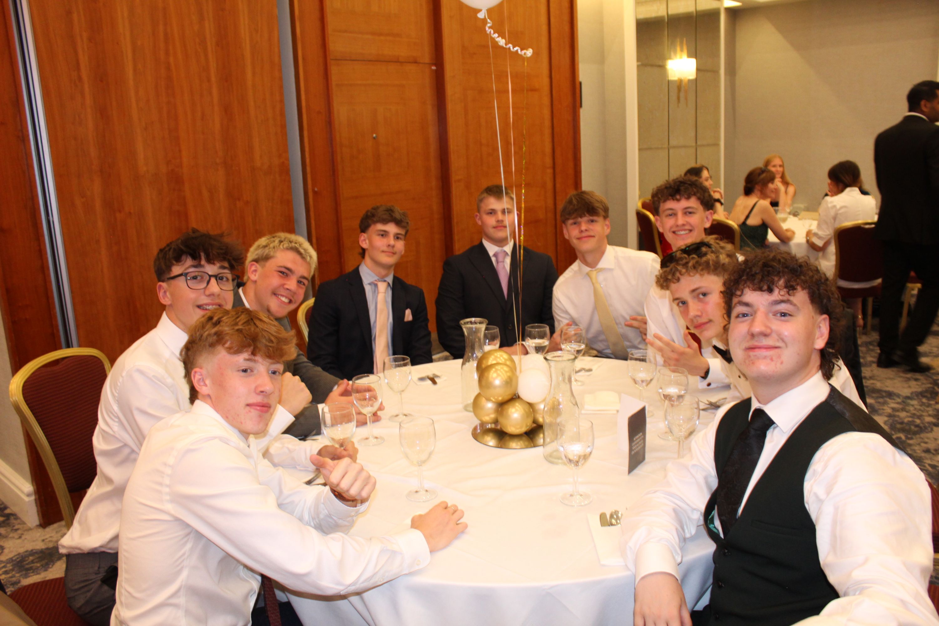 Photograph of a group of nine young men dressed in formal attire seated around a round table at an indoor event. Table features a white tablecloth, empty glasses, and a gold ornament centerpiece with balloons,at prom