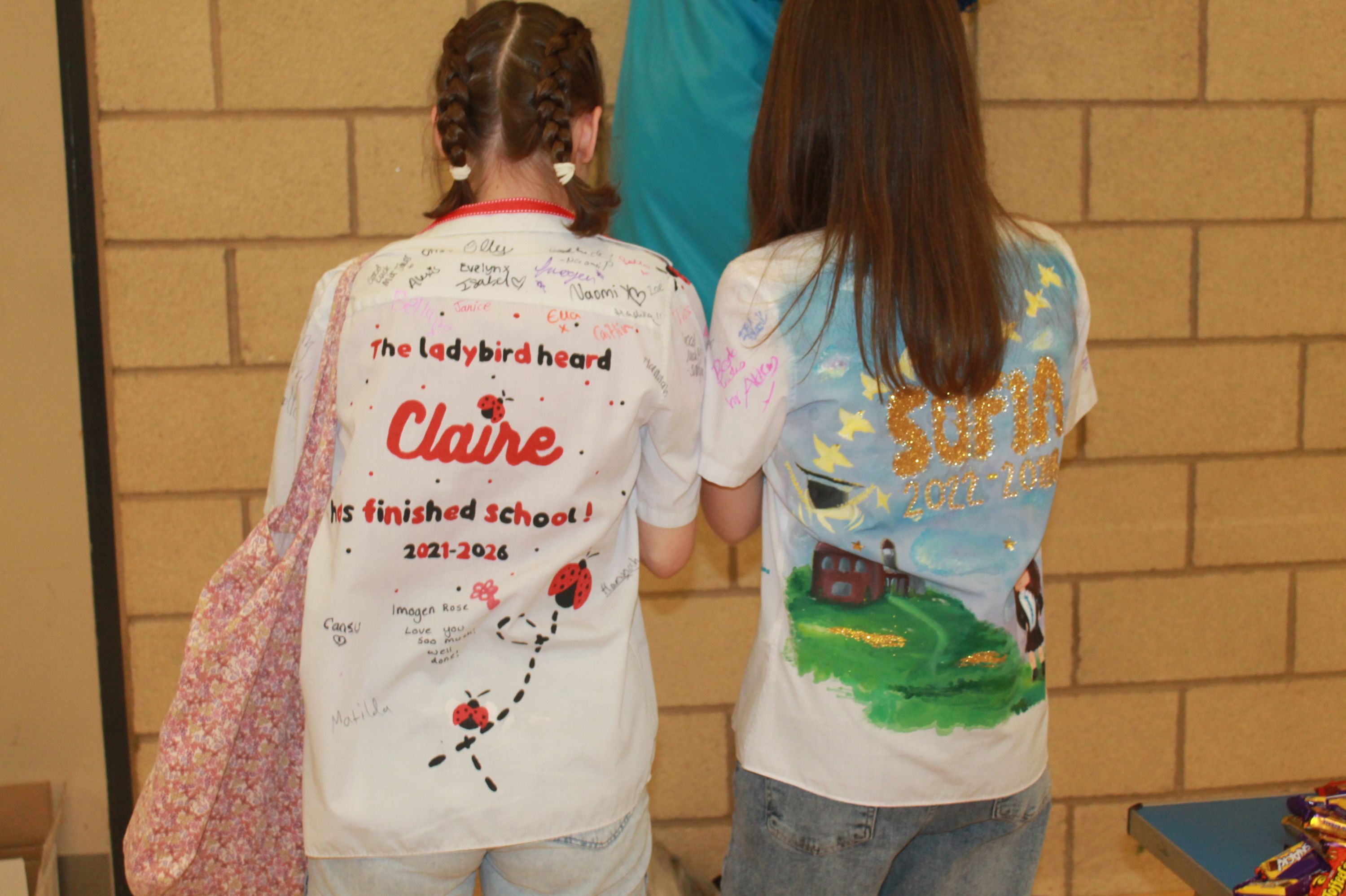 Photograph of two children wearing decorated white T-shirts, standing with backs to camera against a brick wall. Left shirt features red text and ladybird illustrations celebrating school completion, while right shirt displays a painted countryside scene with stars and text in gold.