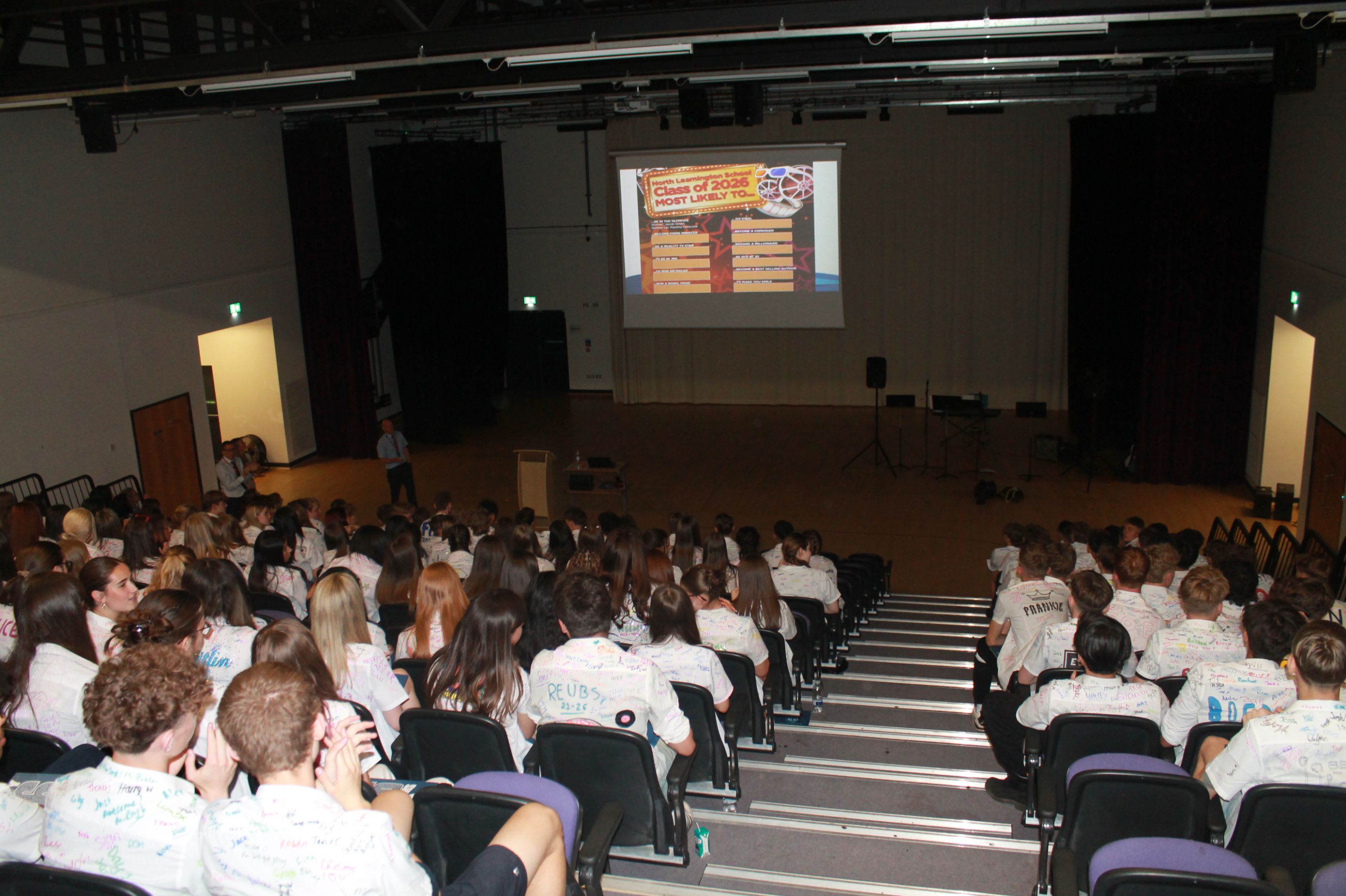 Photograph of an auditorium filled with students wearing white shirts covered in colourful signatures, watching a presentation projected on a screen at front. Presentation slide titled "Class of 2023" lists names or groups in orange and white text with decorative elements, indicating a celebratory or farewell event.
