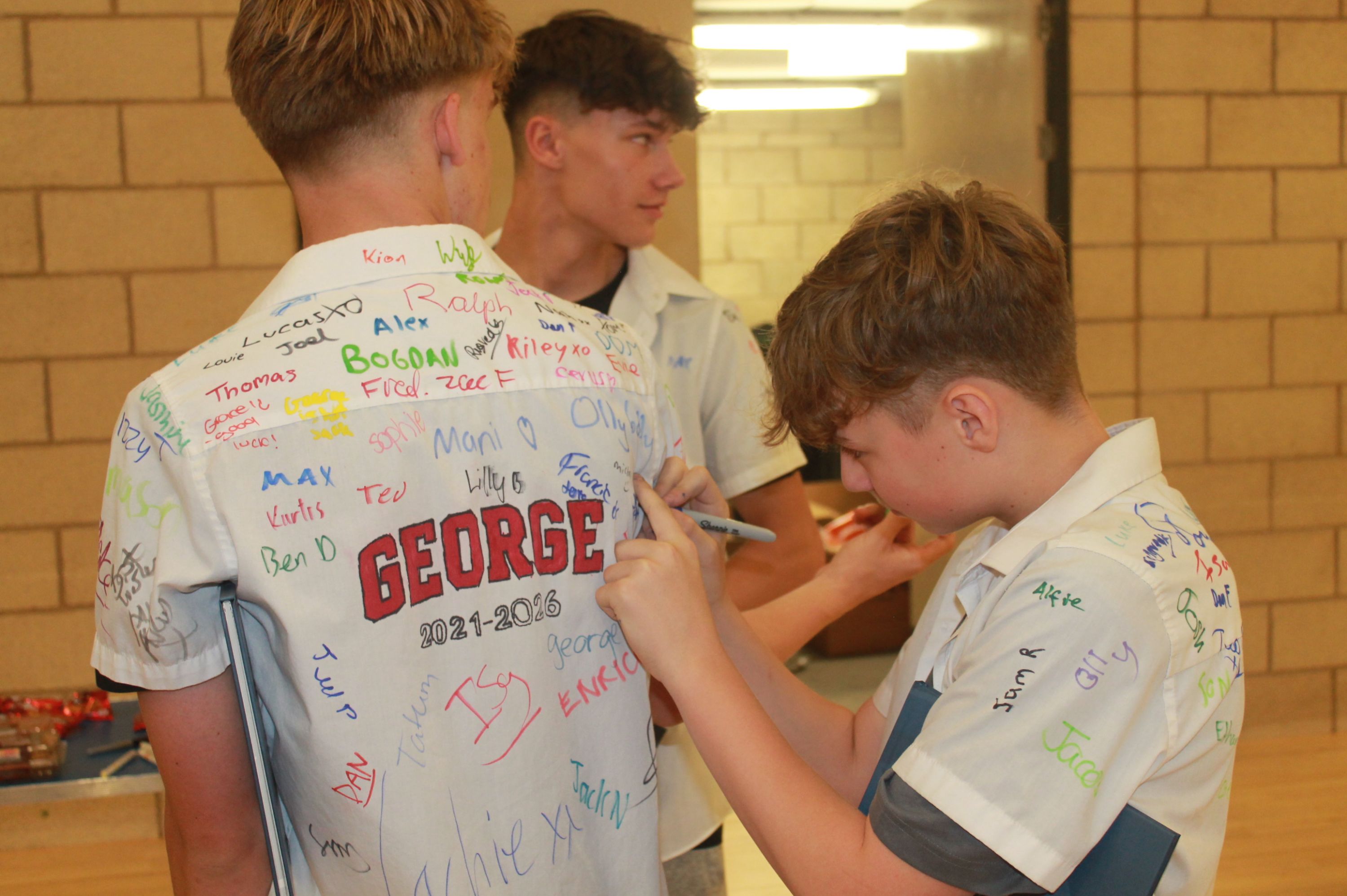 PHotograph showing three boys in a school hall wearing white shirts covered with colourful handwritten signatures and messa