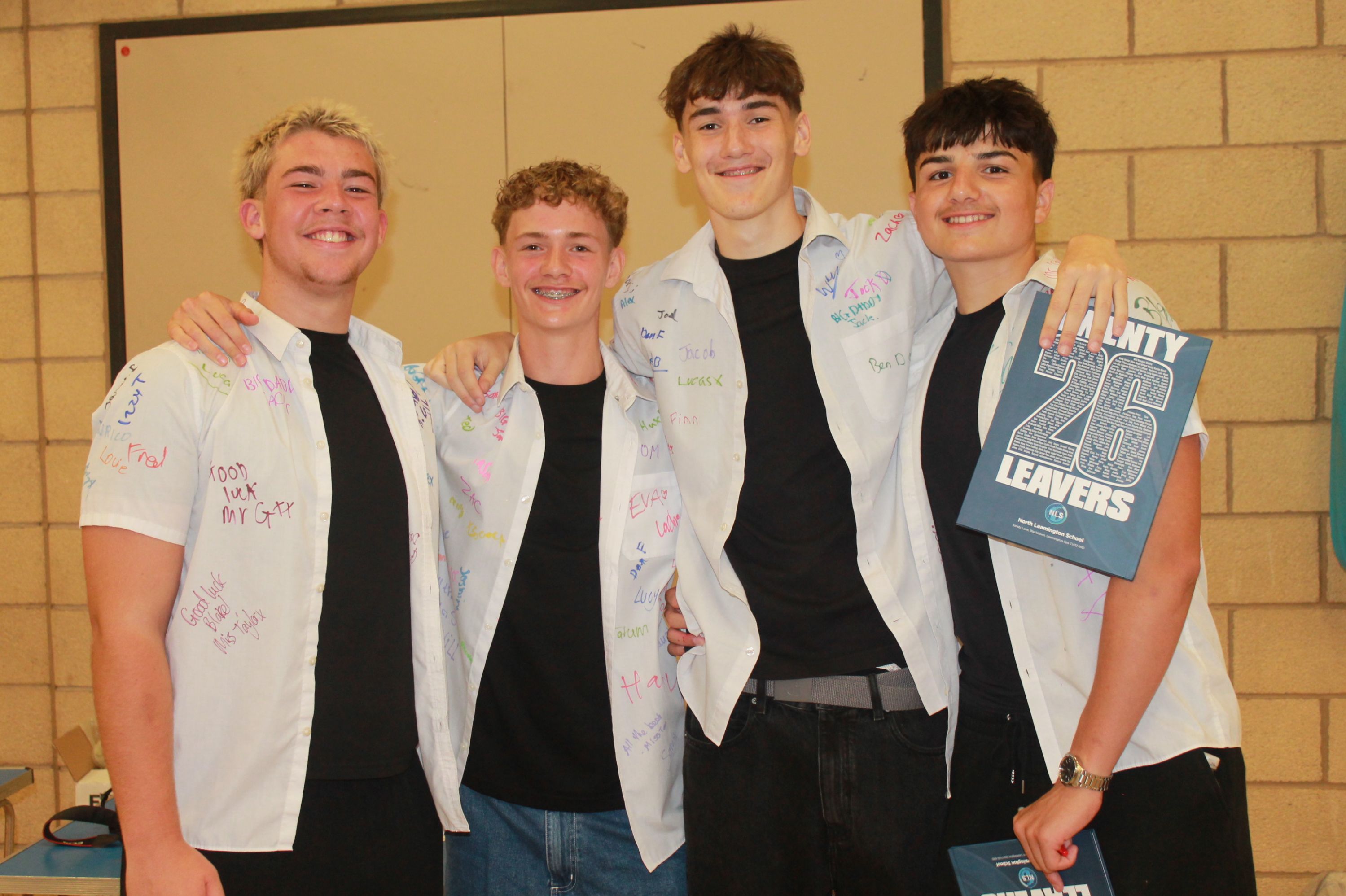 Photograph of four teenage boys standing indoors, smiling and wearing white shirts covered in handwritten messages and signatures. One boy holds a blue book titled "Class of 26 Leavers," indicating a school leaving or graduation event.