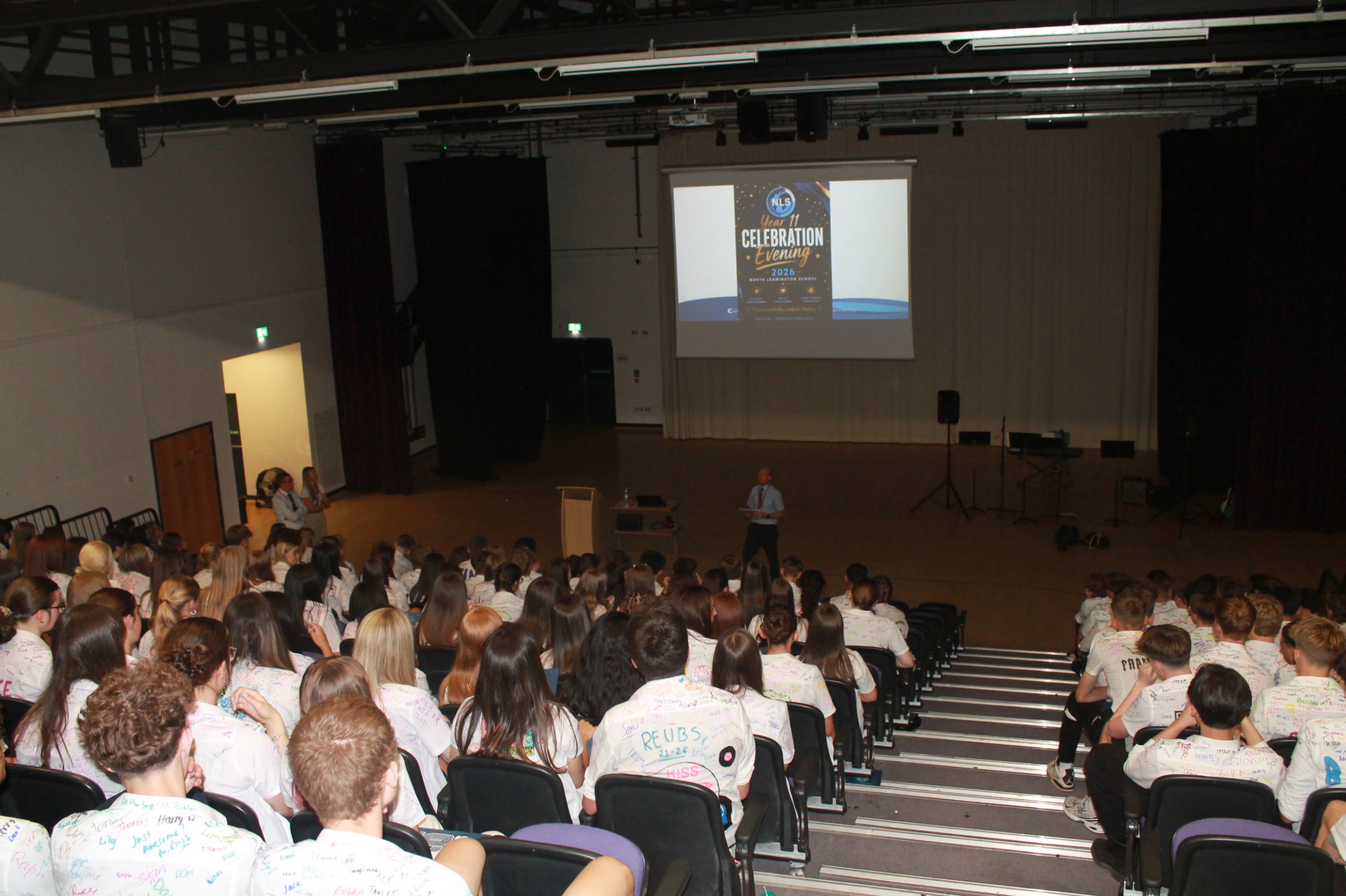 Photograph of a large group of students seated in an auditorium facing a stage with a speaker and a projected presentation titled "Celebration of Learning." Students wear white shirts covered in colourful signatures, indicating a special event or milestone celebration, with the setting highlighting an educational or school environment.