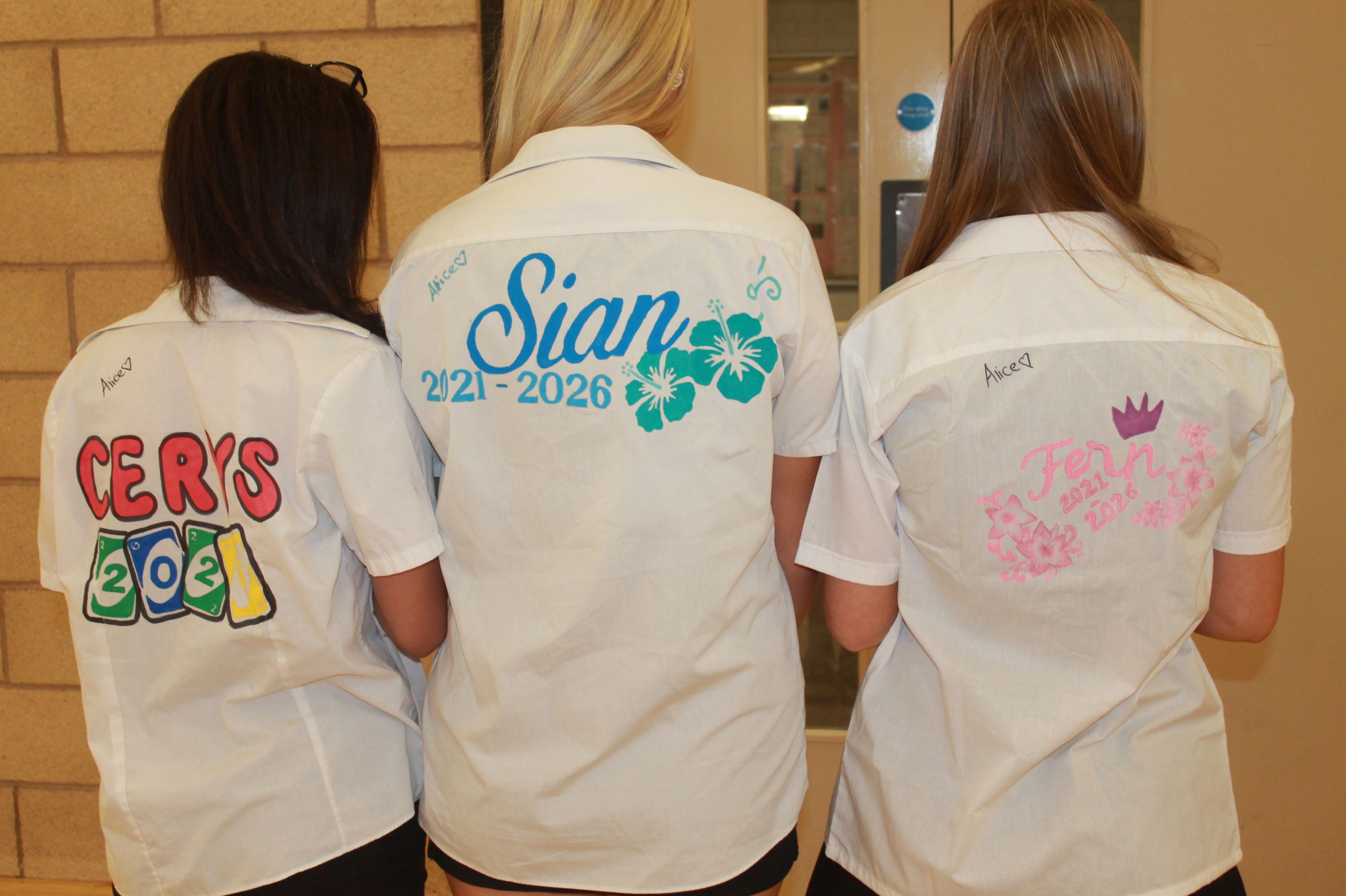 Photograph of three people wearing white shirts customised with names and dates on backs, likely representing school or college leavers. Shirts feature colourful designs including bold text, floral patterns, and graduation caps, with names "Cerys," "Sian," and "Fern" and years "2021," "2021-2026," and "2021-2026" 
