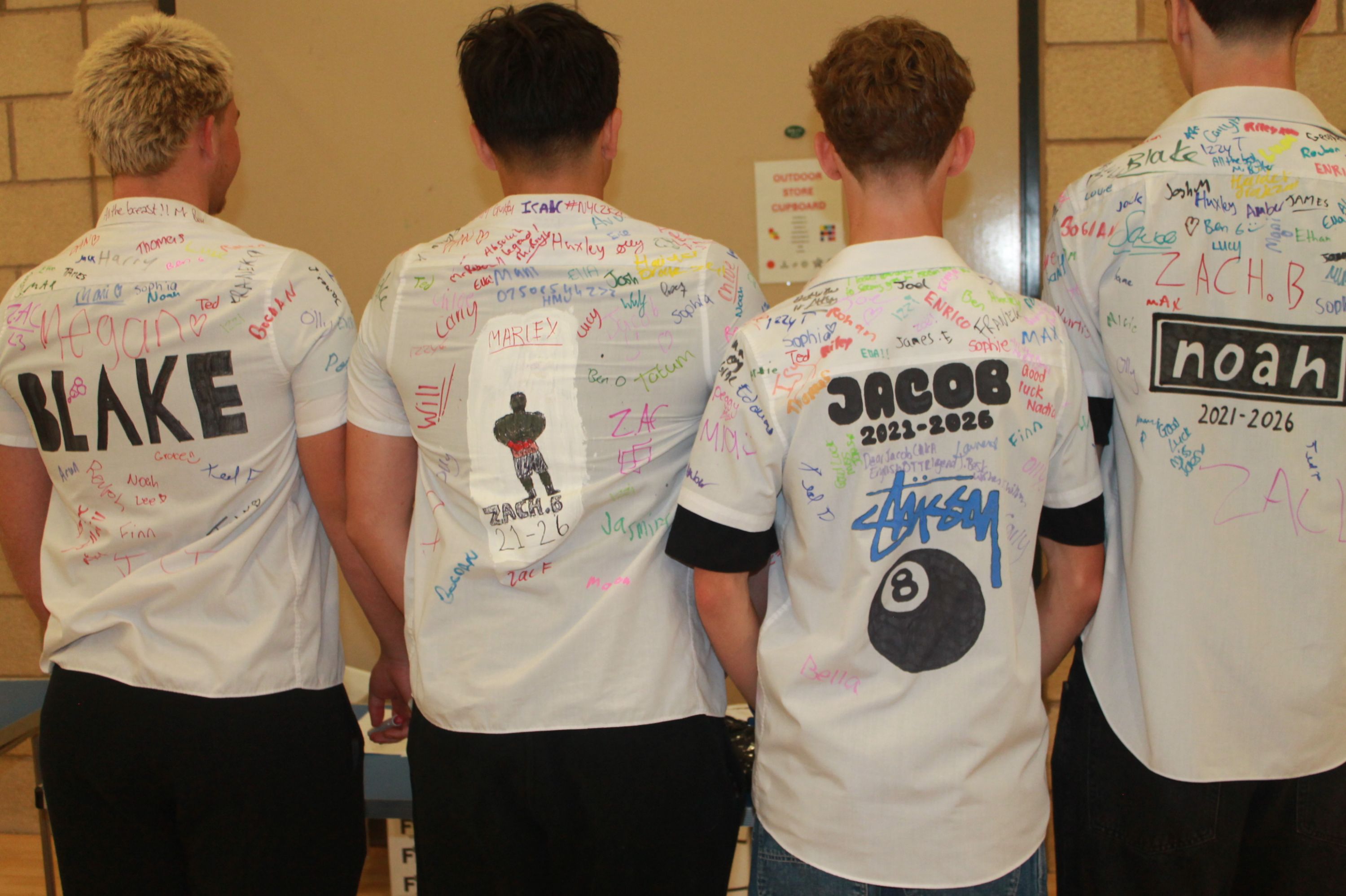 Photograph showing four students from behind wearing white shirts signed with colourful markers, indicating a celebration or farewell event. Each shirt features a large name label and personalised designs, such as a silhouette, an eight ball, and date ranges from 2021 to 2026, highlighting individual identities and memories.