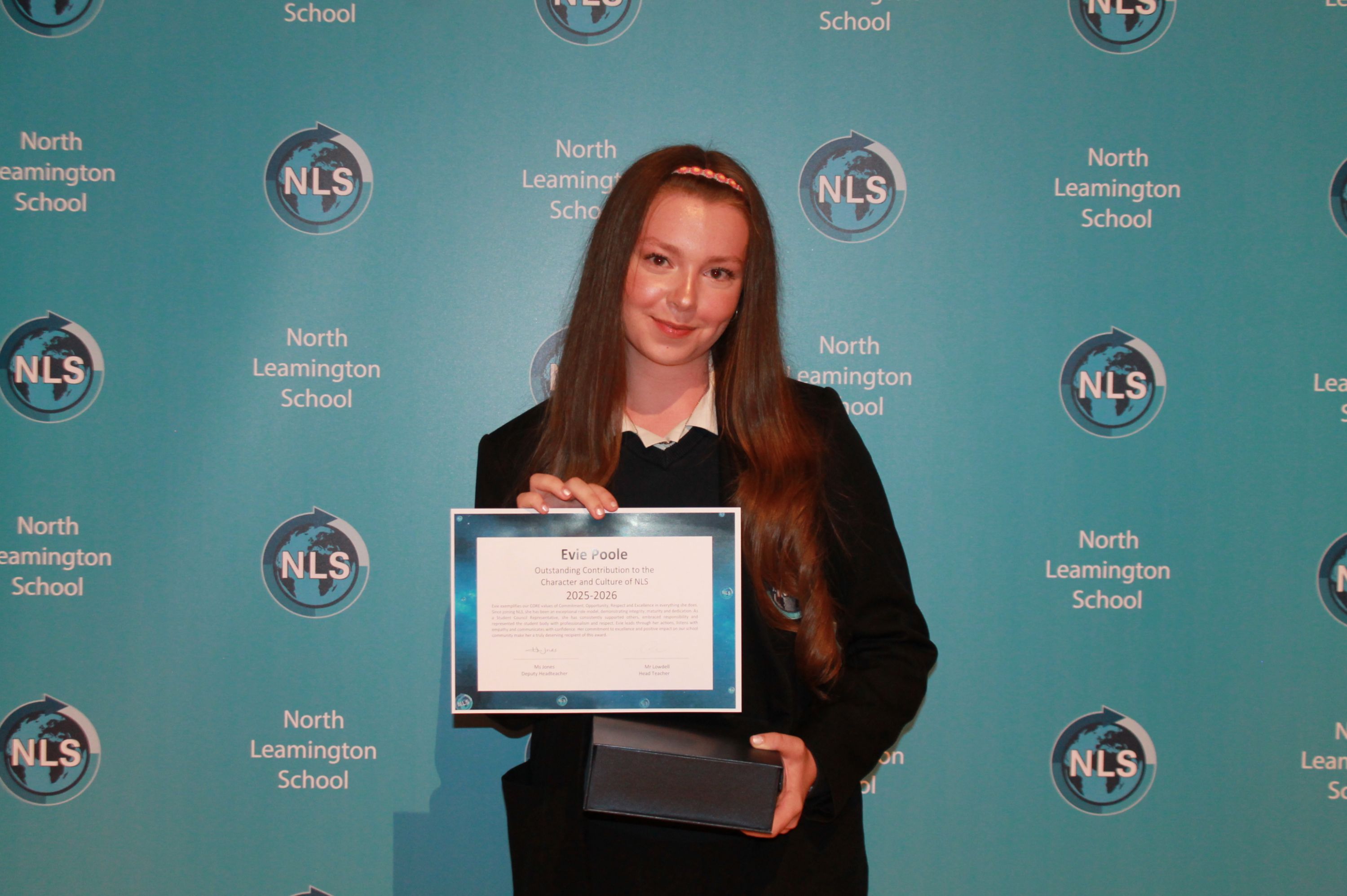Photograph of a young woman standing in front of a North Leamington School backdrop holding a certificate and a small box, likely during an award or graduation ceremony. The backdrop features repeated school logos and text in white on a teal background, highlighting the school's branding.