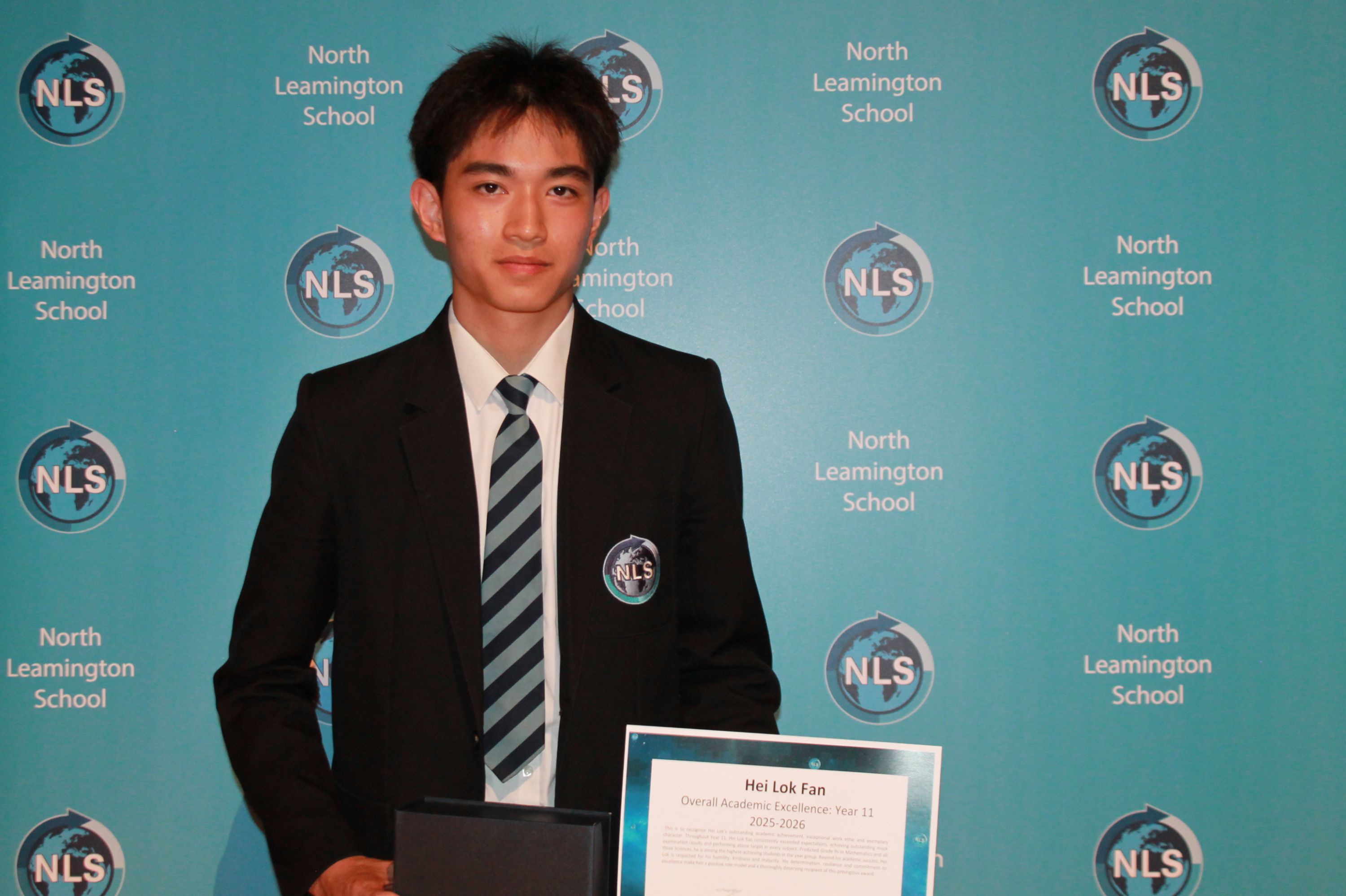 Photograph of a young male student in a school uniform holding an award certificate and a box, standing in front of a blue backdrop with repeated "North Leamington School" and "NLS" logos. The award certificate recognises academic excellence for Year 11