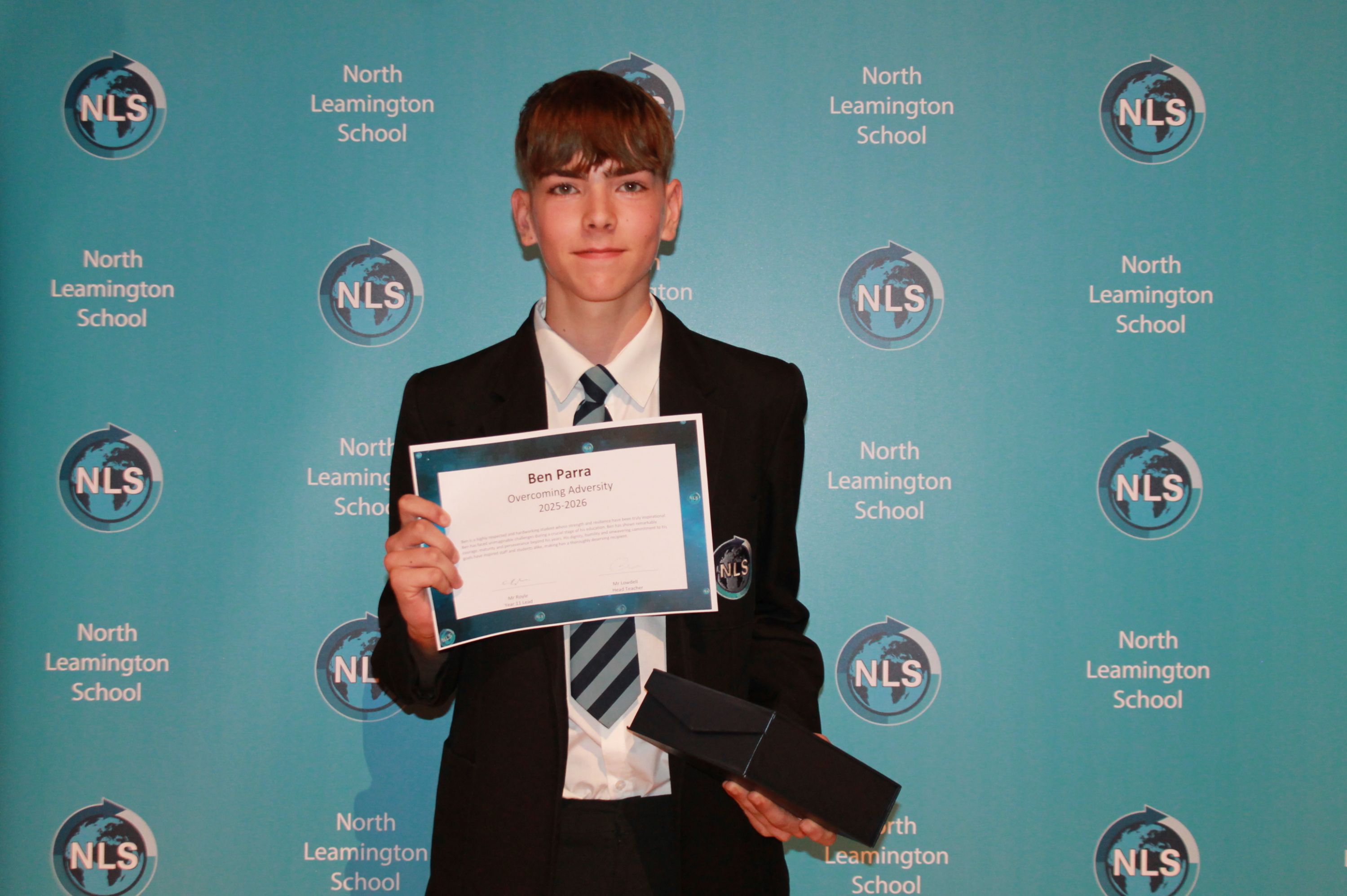 Photograph of a young male student in school uniform holding a certificate and a black box, standing in front of a blue backdrop with repeated "North Leamington School" and "NLS" logos. 