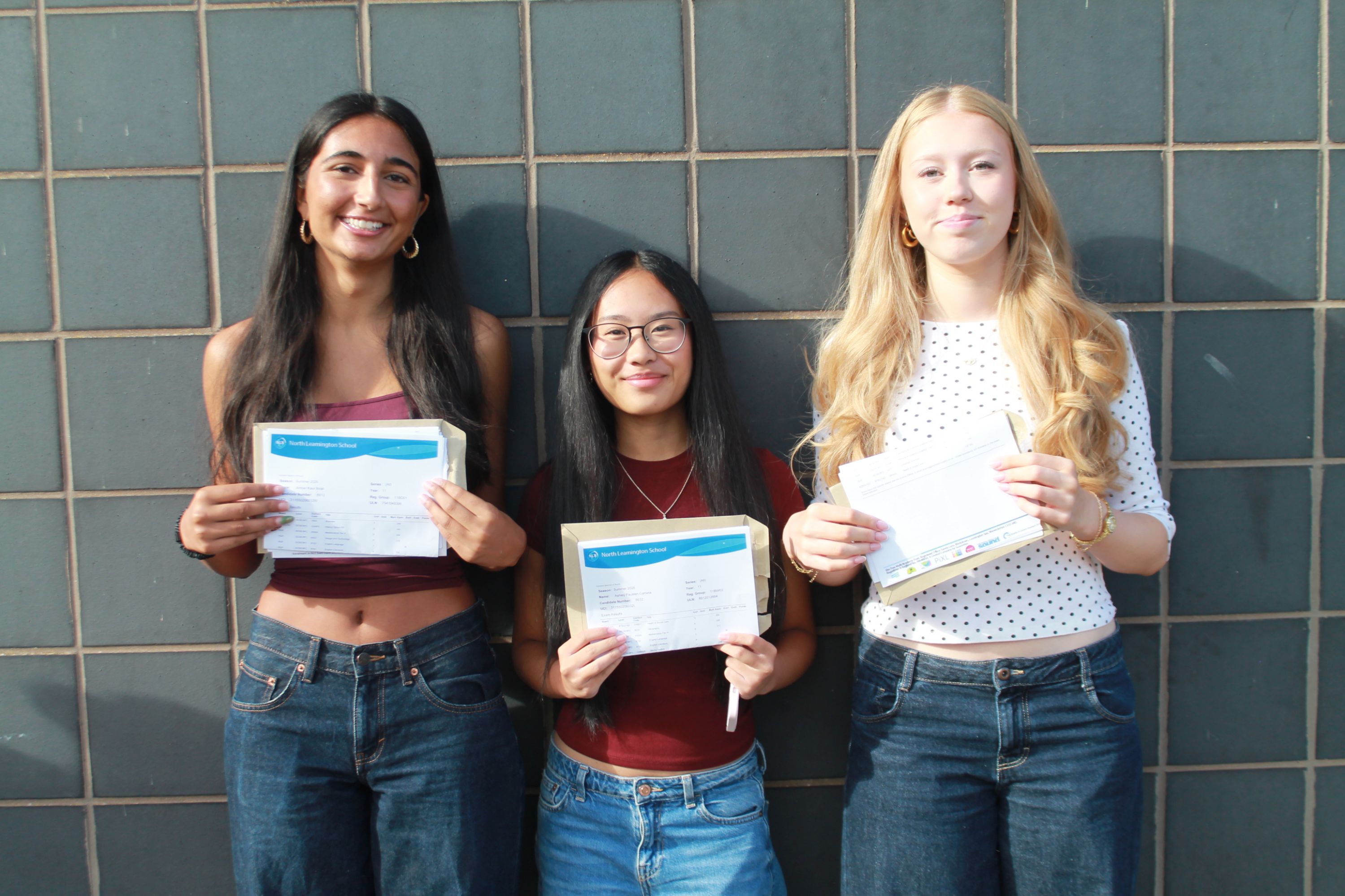 Photograph of three young women standing side by side against a tiled wall, each holding documents or envelopes