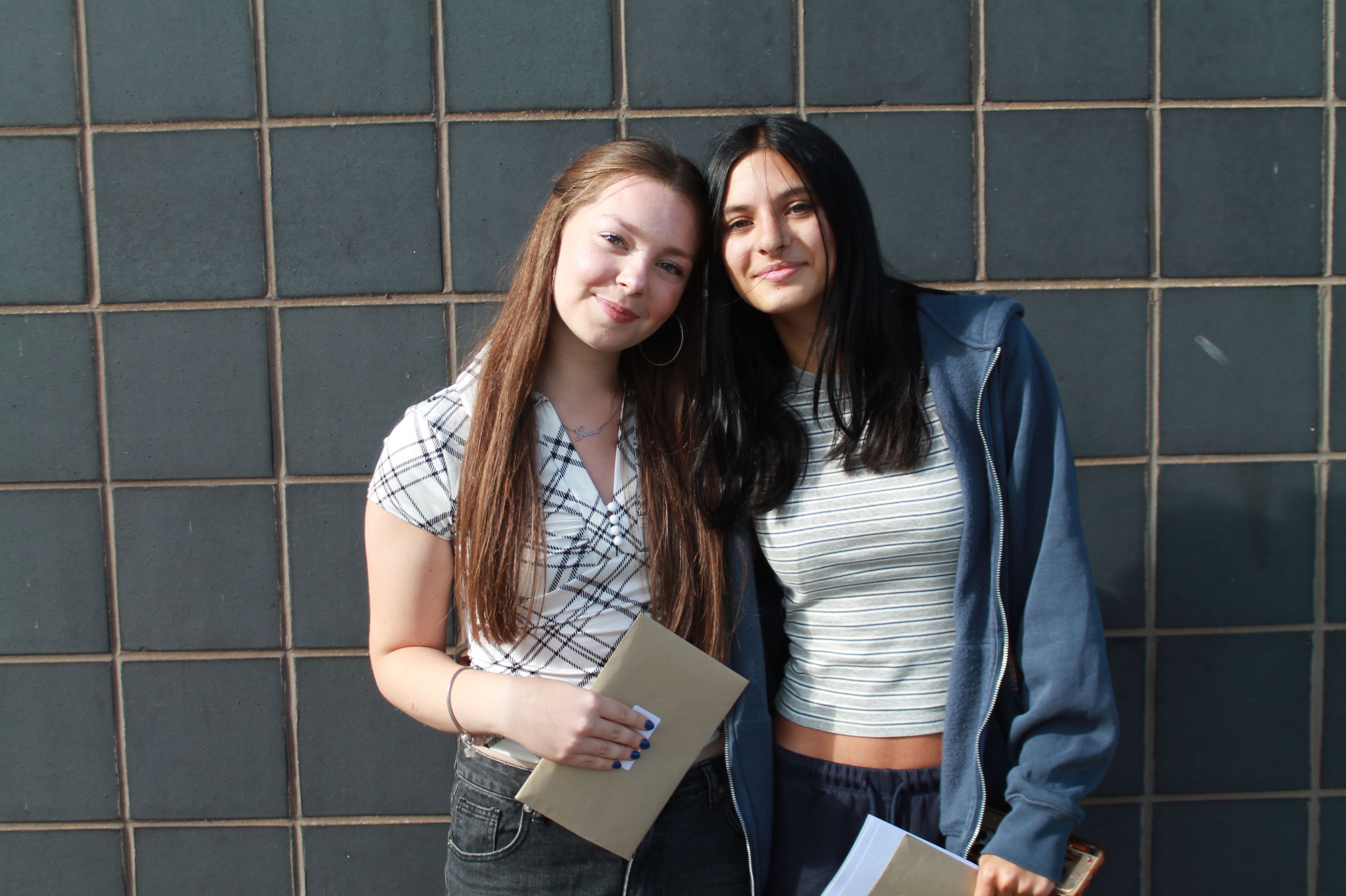 Photograph of two young women standing side by side against a tiled wall, each holding documents or envelopes