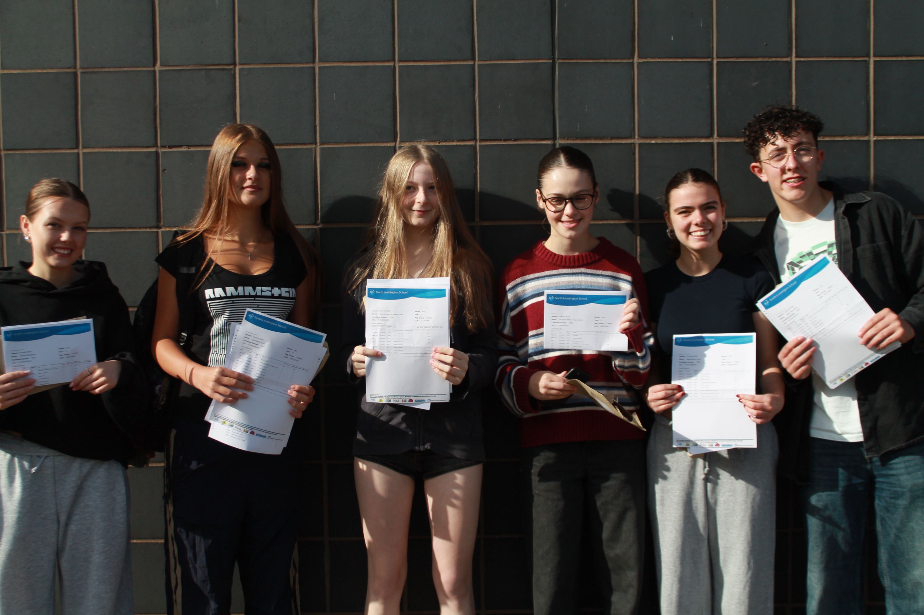 Photograph of six young people standing side by side against a tiled wall, each holding documents or envelopes