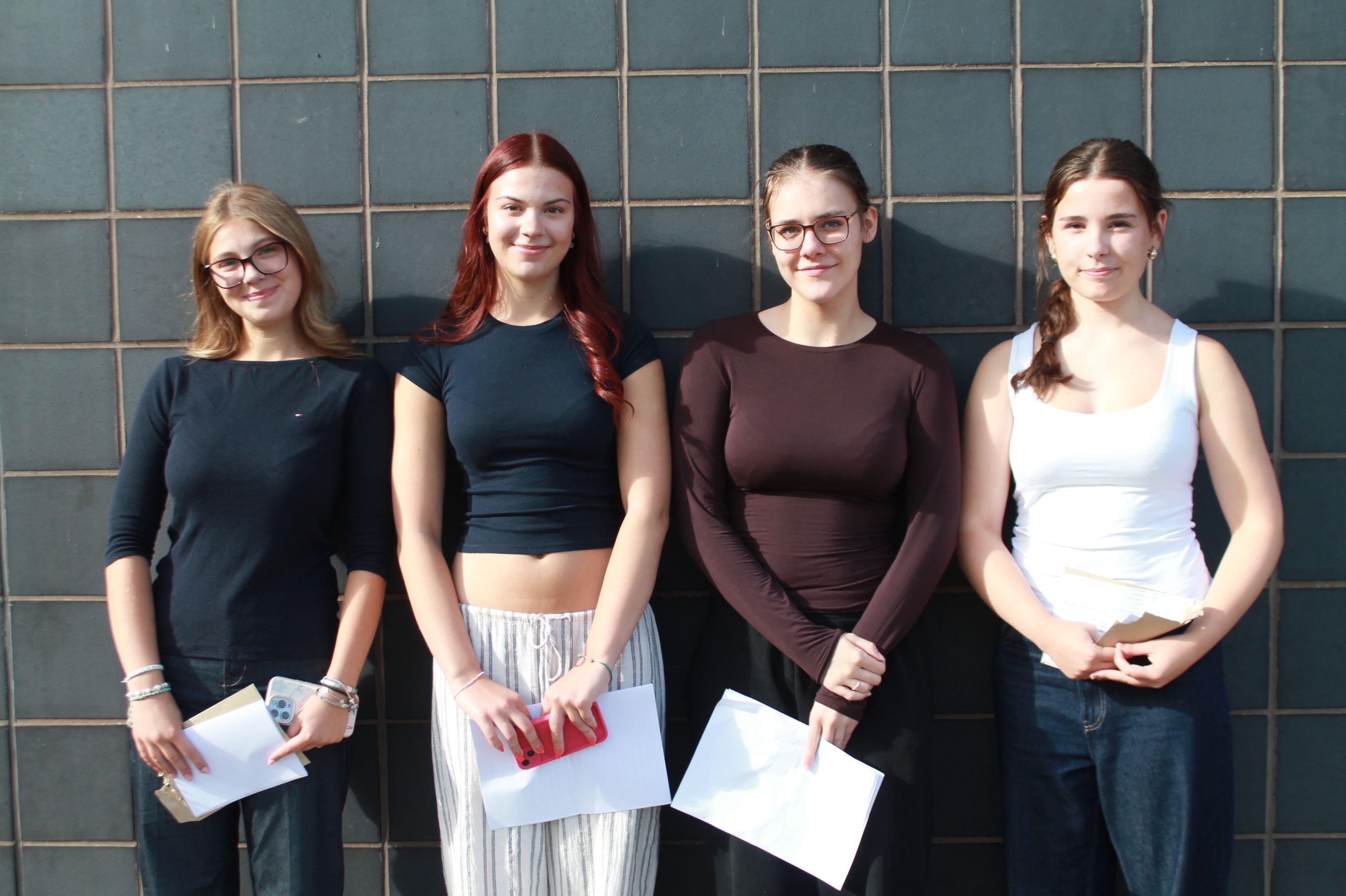 Photograph of four  young women standing side by side against a tiled wall, each holding documents or envelopes