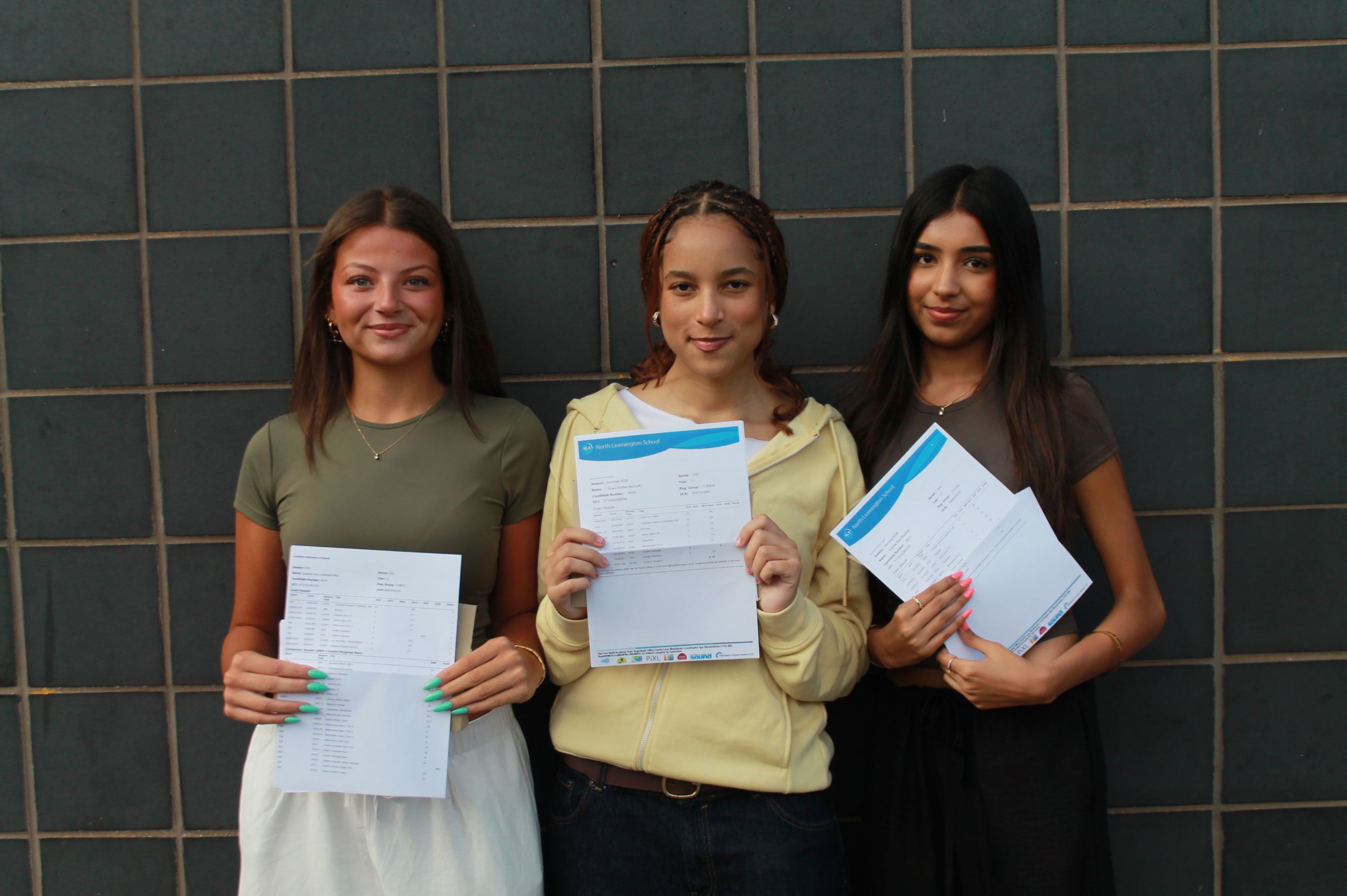 Photograph of three young women standing side by side against a tiled wall, each holding documents or envelopes