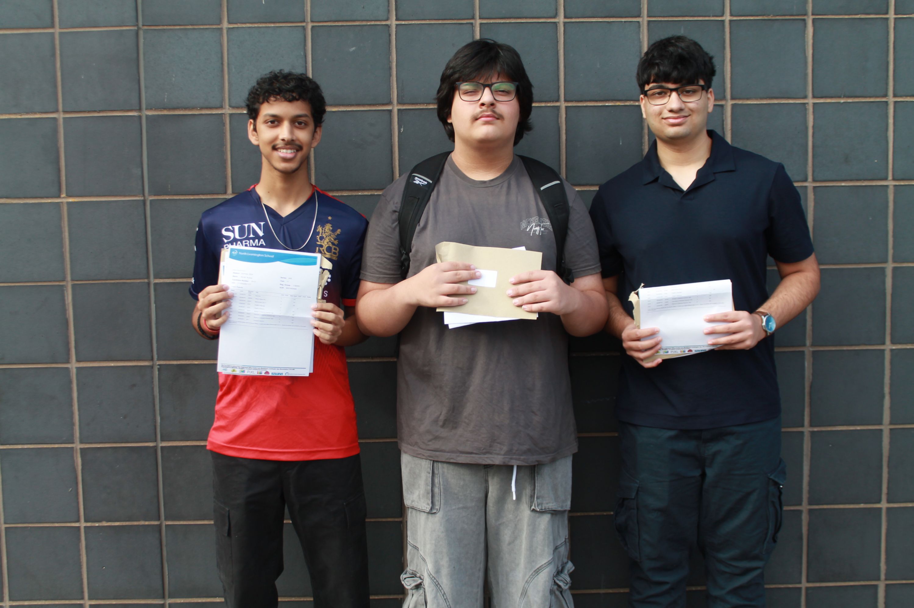 Photograph of three young men standing side by side against a tiled wall, each holding documents or envelopes
